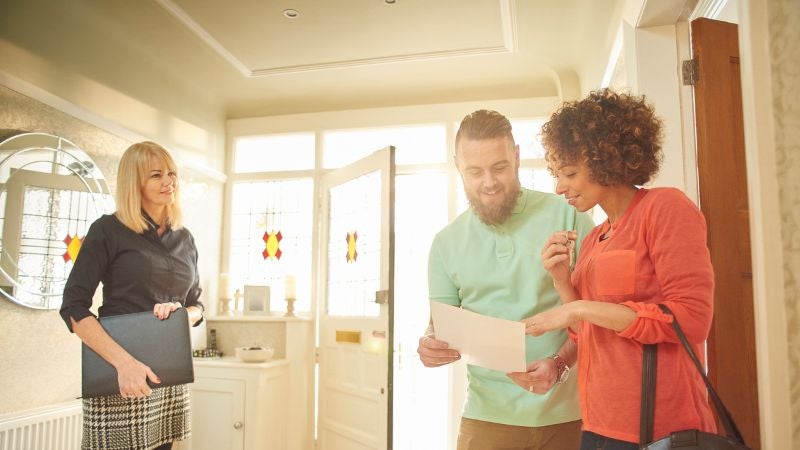 Real estate agent smiles at couple looking at piece of paper inside of a brightly-lit house.