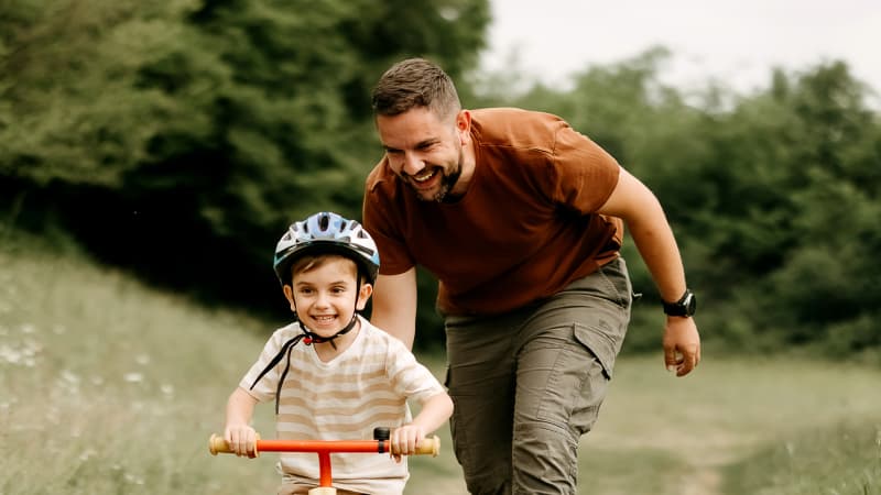 Father in a park supporting his young son who is learning to ride a bike.